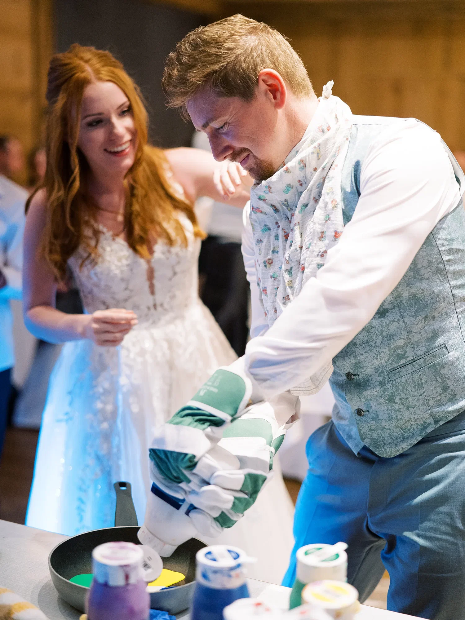Der Bräutigam trägt Handschuhe und ist mitten in einem lustigen Hochzeitsspiel, bei dem er mit Farbe hantiert, die Braut steht daneben und lächelt.