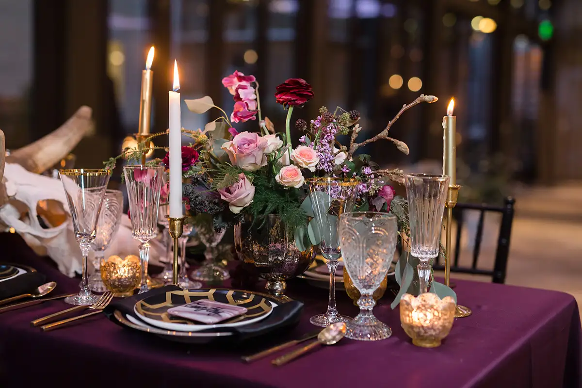 Ein elegant gedeckter Tisch mit goldverzierten Weingläsern, brennenden Kerzen und einem Blumengesteck aus rosa und roten Blüten, arrangiert in einem stilvollen, dunklen Setting.