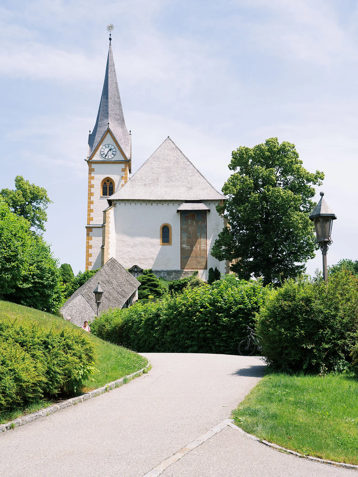 Historische Kirche mit spitzem Turm und Uhr, umgeben von Bäumen und Büschen, an einem sonnigen Tag mit klarem Himmel.