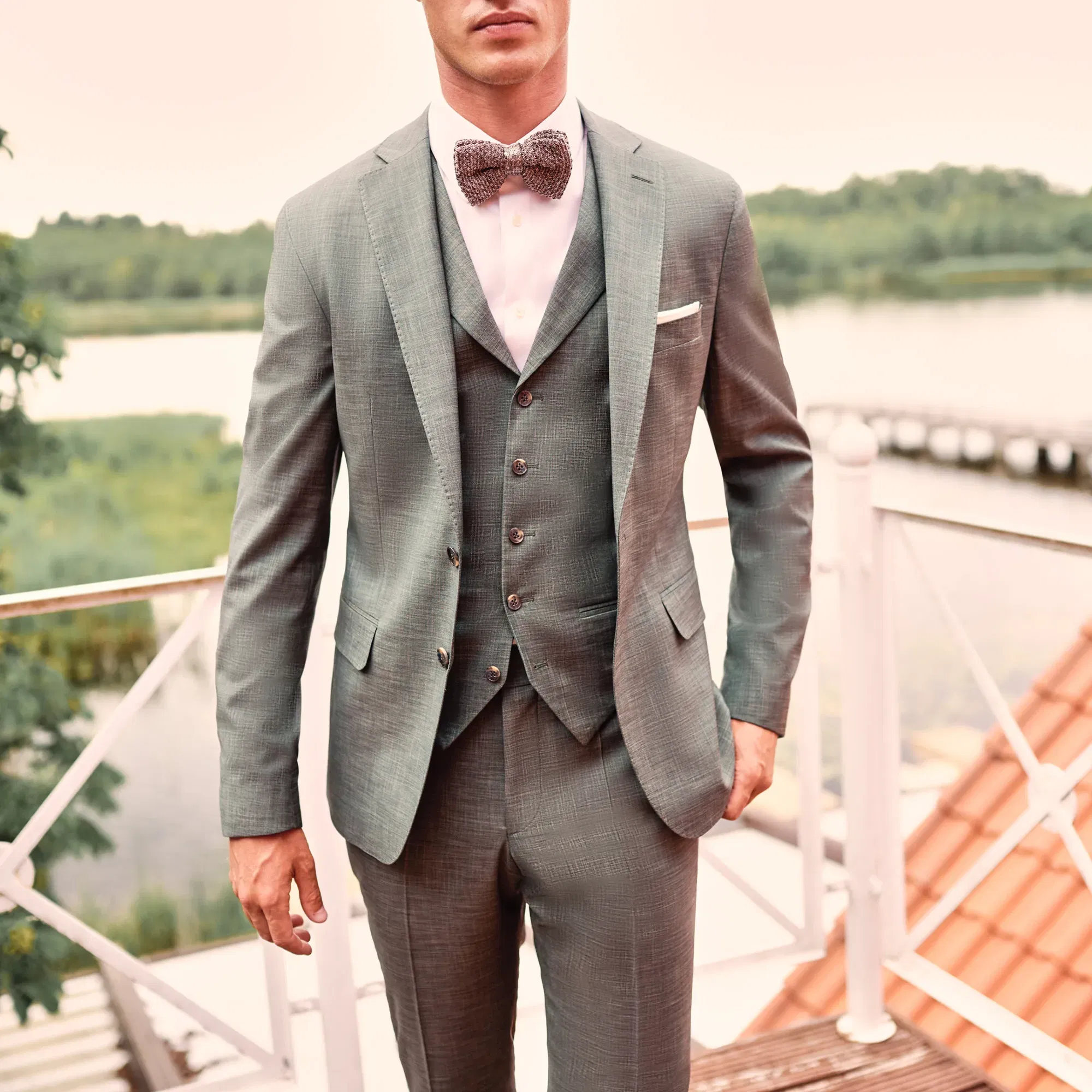 Ein Bräutigam in einem stilvollen, grauen Anzug mit passender Weste und Fliege posiert selbstbewusst auf einer Terrasse mit Blick auf eine ruhige Landschaft.