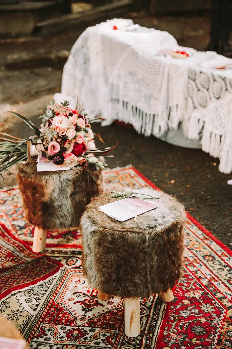 Ein flauschiger Fellhocker steht auf einem bunten Teppich, daneben liegt ein eleganter Blumenstrauß mit Rosen und grünen Blättern. Der zeremonielle Tisch im Hintergrund ist mit einer gehäkelten Decke dekoriert, was eine gemütliche Atmosphäre schafft.