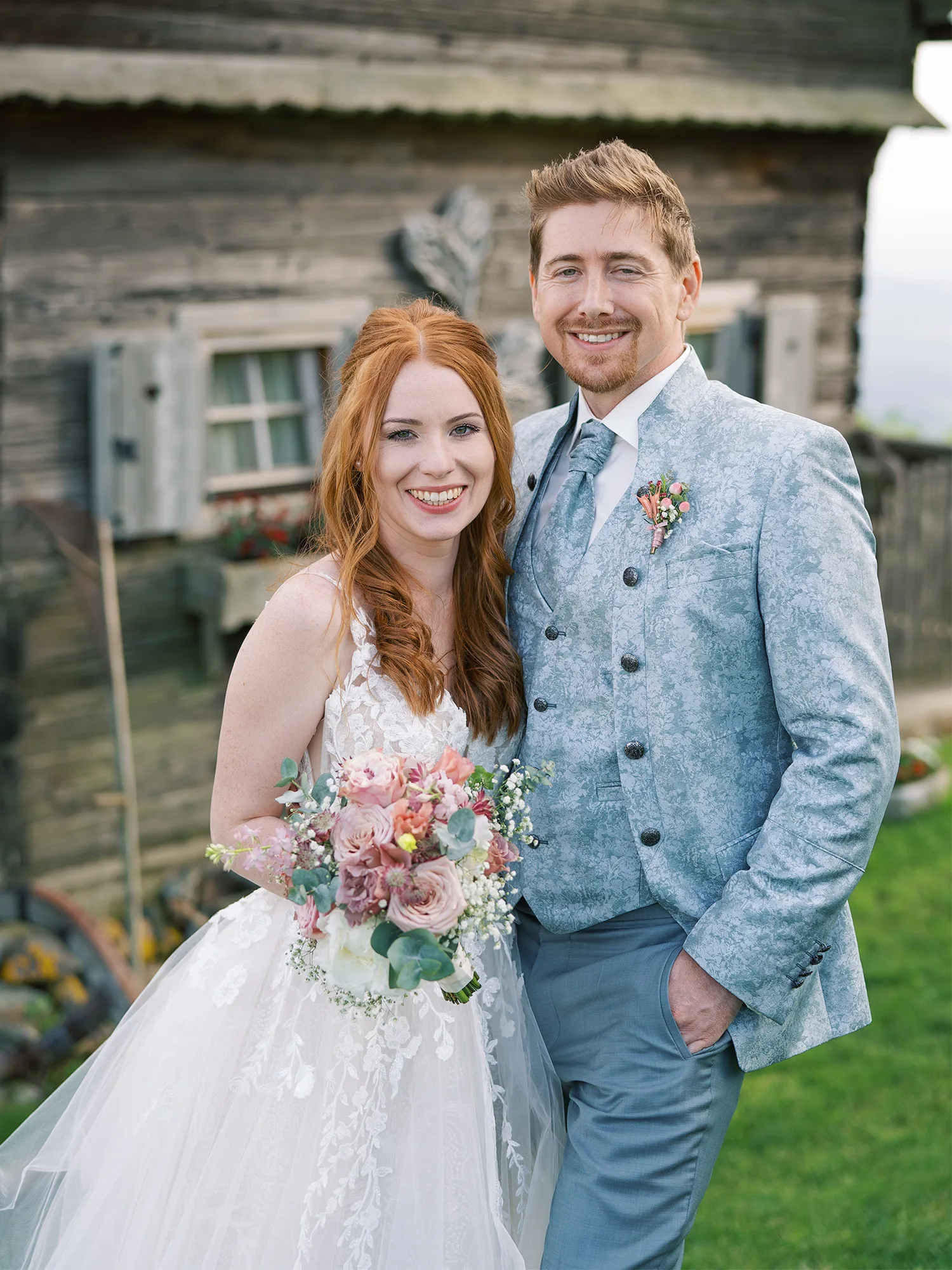 Braut und Bräutigam posieren vor einem alten Holzhaus, sie trägt ein weißes Spitzenkleid und hält einen romantischen Brautstrauß, während er in einem hellgrauen Anzug lächelt.