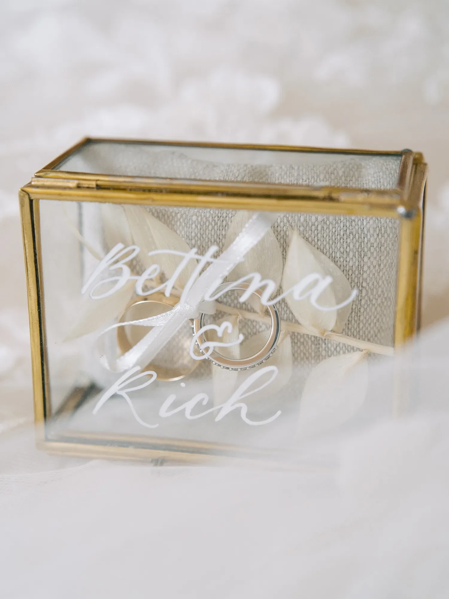 Nahaufnahme von Eheringen in einer goldenen Glasbox mit der Aufschrift "Bettina & Rich", umgeben von zarter Dekoration.