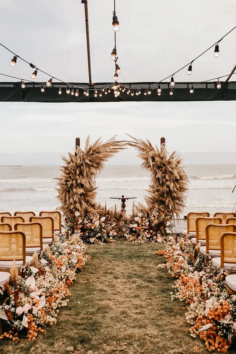 Eine romantische Hochzeitsszene am Meer mit einem imposanten Pampasgrasbogen und üppigen Blumenarrangements entlang des Gangs. Die Stühle sind in Richtung des Altars ausgerichtet, mit dem Ozean als friedliche Kulisse im Hintergrund.