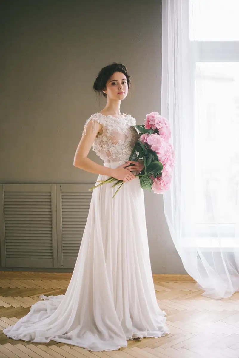 Die Braut, gekleidet in einem zarten weißen Kleid, trägt einen voluminösen Strauß rosafarbener Pfingstrosen. Sie steht in einem schlicht eingerichteten Raum.