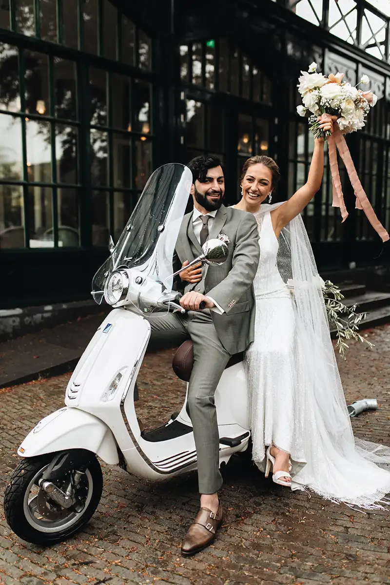 Ein frisch verheiratetes Paar, das auf einer Vespa posiert, die Braut jubelt mit einem Blumenstrauß, während der Bräutigam lächelnd die Vespa lenkt.