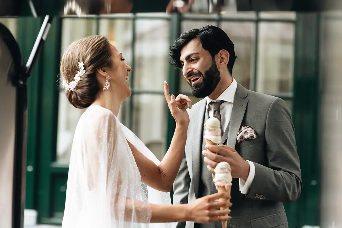 Braut und Bräutigam, die Eiscreme genießen und sich gegenseitig necken, während sie stilvolle Hochzeitskleidung tragen und fröhlich miteinander interagieren.
