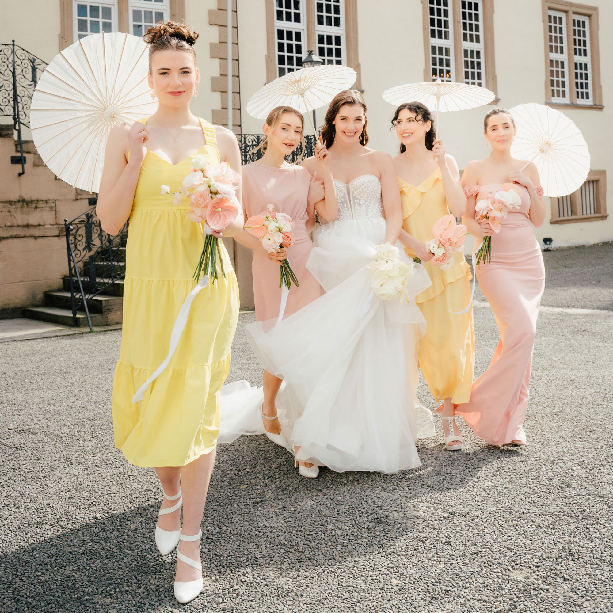 Eine Braut in einem weißen Hochzeitskleid steht mit ihren vier Brautjungfern, die in Gelb und Rosa gekleidet sind, vor einem klassischen Herrenhaus. Die Brautjungfern tragen Blumengestecke und halten weiße Sonnenschirme, die eine sommerliche Atmosphäre schaffen. Die Gruppe lächelt und posiert für die Kamera, während die Sonne den Tag erhellt.