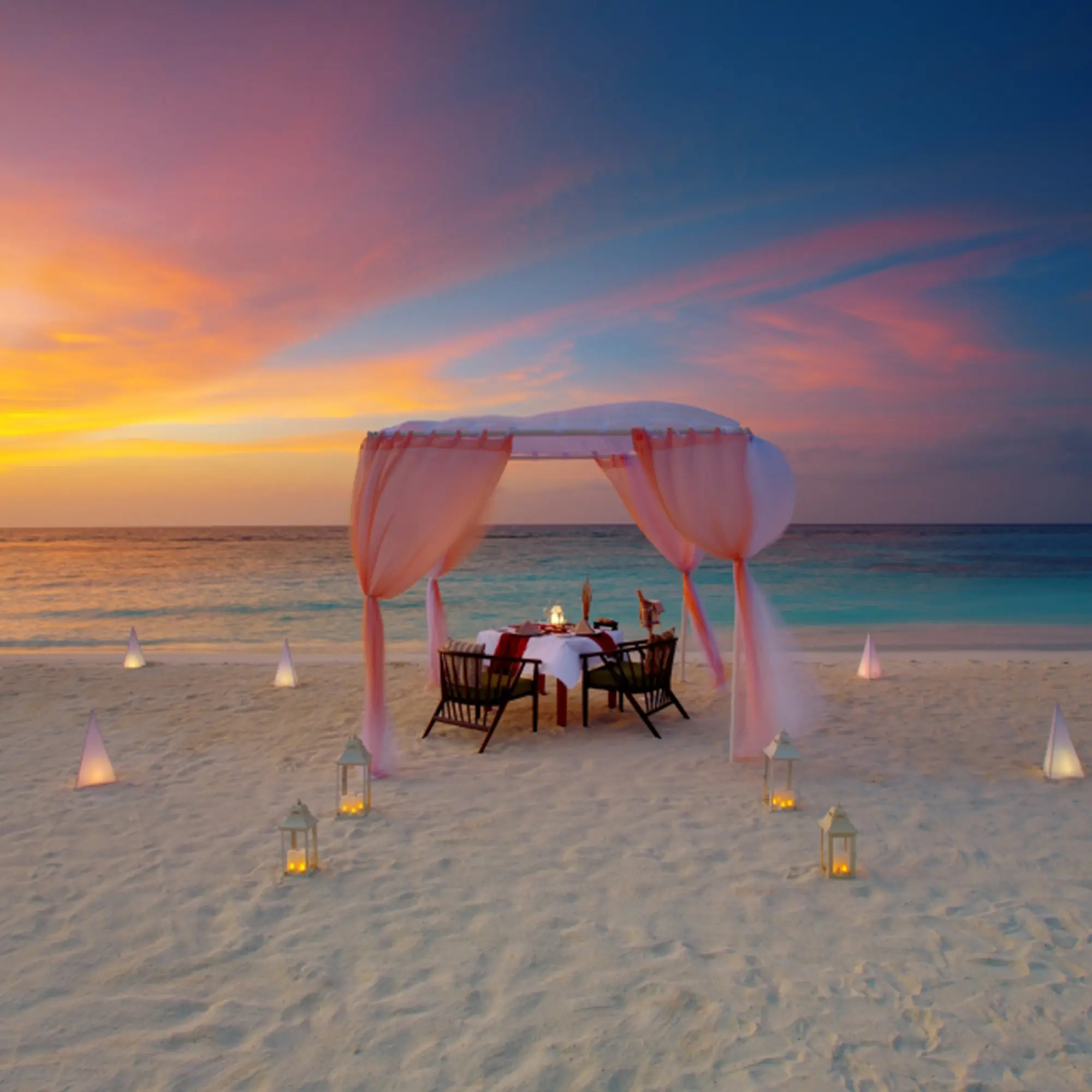 Privates romantisches Dinner am Strand mit einem elegant gedeckten Tisch unter einem Stoffbaldachin, während die Sonne am Horizont untergeht.