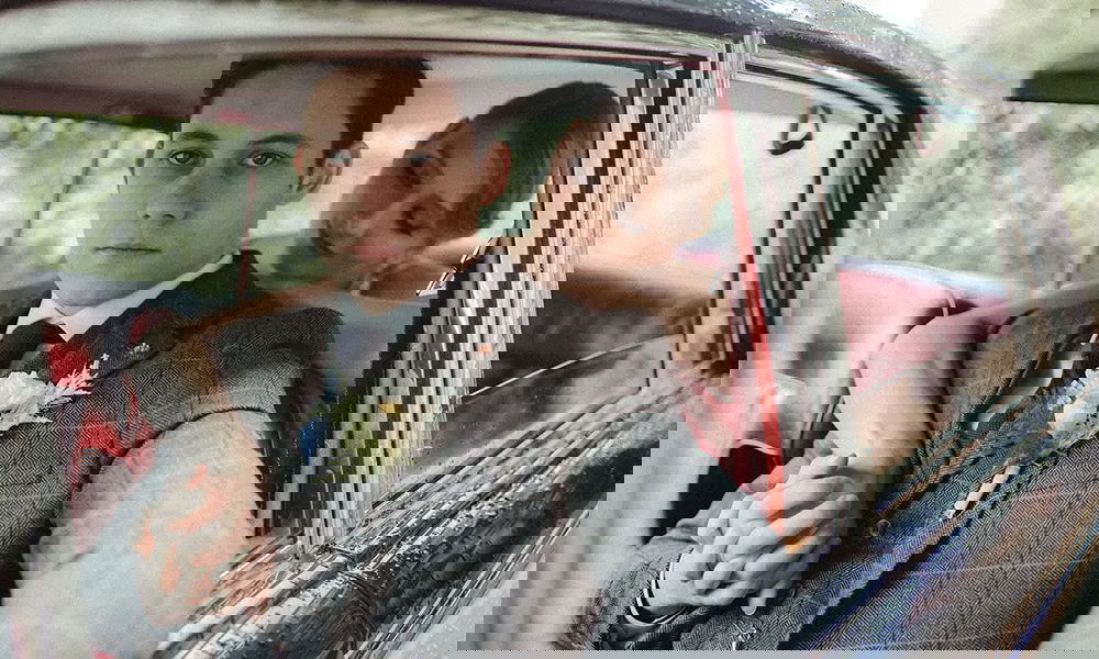 Ein frisch verheiratetes Paar in einem eleganten Oldtimer: Der Bräutigam in einem klassischen grauen Anzug mit Krawatte blickt ernst in die Kamera, während die Braut sich mit einem sanften Lächeln an ihn schmiegt. Der Wagen mit roter Lederausstattung und regennassen Fenstern verstärkt die nostalgische Atmosphäre.