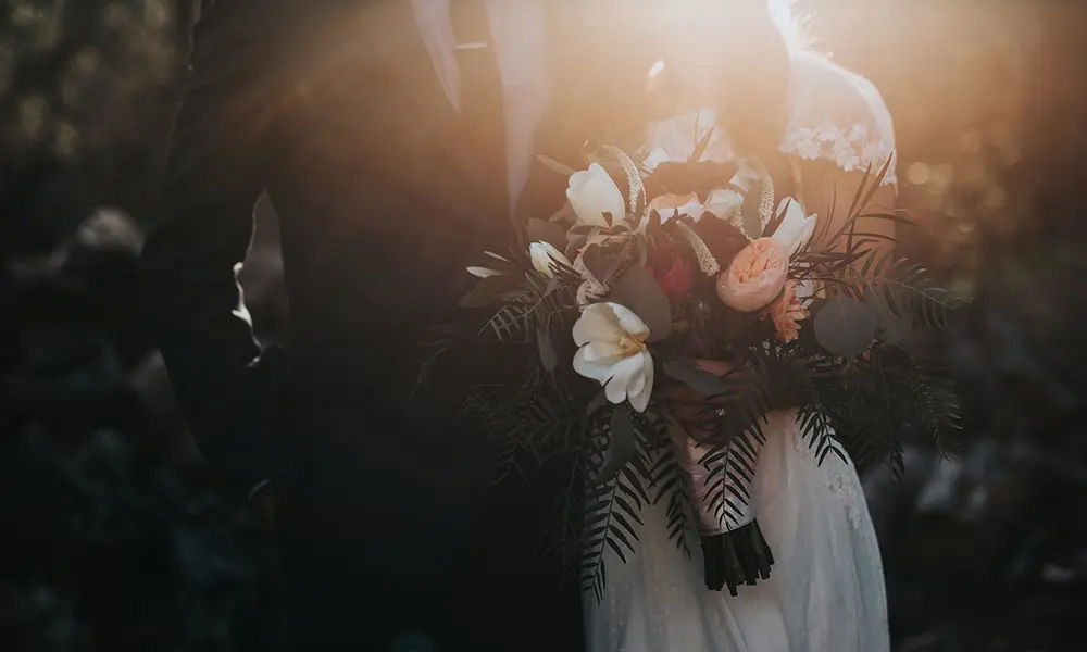 Stimmungsvolles Hochzeitsfoto bei Sonnenuntergang: Die Braut hält einen üppigen Strauß aus weißen und rosa Blumen mit viel Grün, während das Licht der untergehenden Sonne die romantische Szene umrahmt. Der Bräutigam steht in einem klassischen Anzug an ihrer Seite.