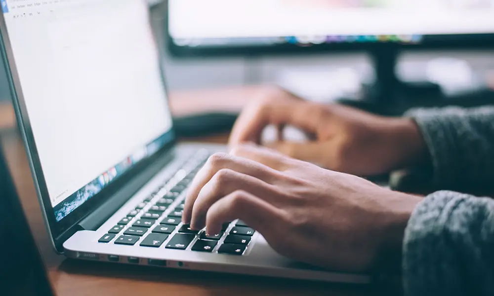 Nahaufnahme von Händen, die auf einem Laptop-Keyboard tippen. Im Hintergrund sind leicht verschwommene Monitore zu sehen, die eine Arbeitsumgebung andeuten.