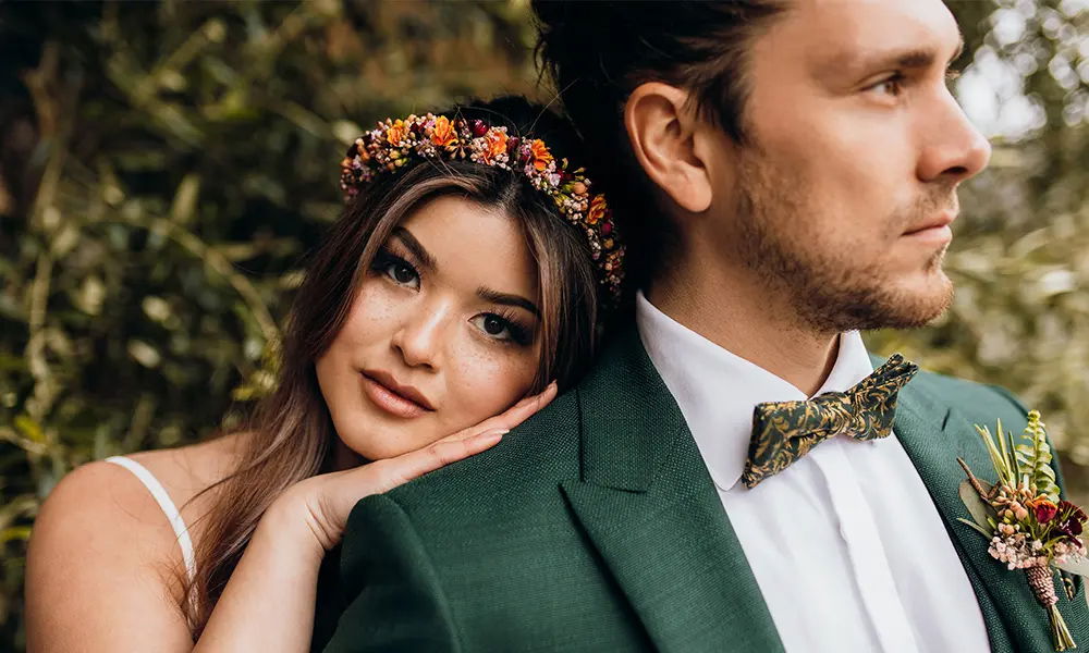 Ein Brautpaar in romantischer Pose: Die Braut mit Blumenkranz lehnt ihren Kopf sanft auf die Schulter des Bräutigams, der in einem grünen Anzug mit floraler Fliege gekleidet ist.