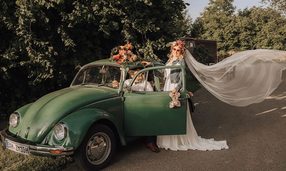 Ein Brautpaar mit Vintage-Flair posiert vor einem grünen VW Käfer. Die Braut trägt ein Hochzeitskleid mit langem Schleier, der im Wind weht, während der Bräutigam im Auto sitzt.