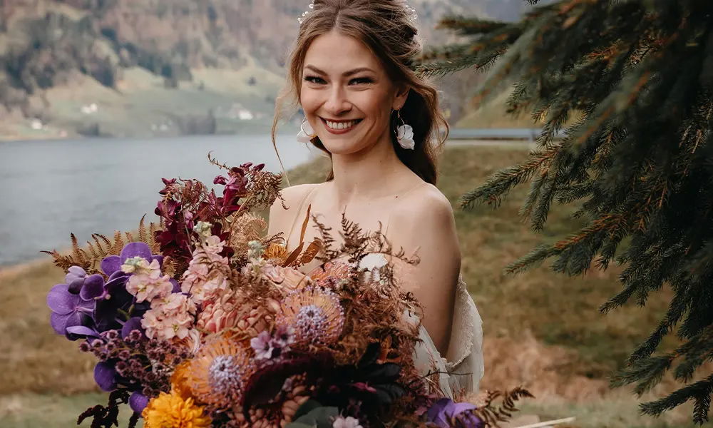 Strahlende Braut in der Natur: Sie hält einen großen, herbstlichen Blumenstrauß mit lila, rosafarbenen und goldenen Akzenten, während der Hintergrund eine ruhige Seelandschaft zeigt.