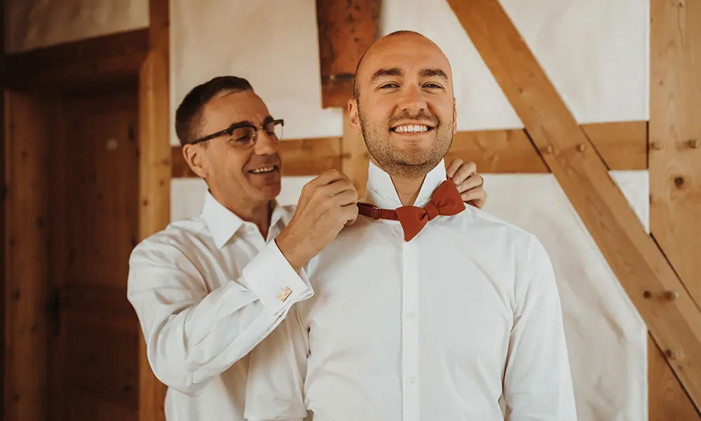 Vorbereitungen vor der Hochzeit: Ein Mann hilft dem Bräutigam, seine rote Fliege zu richten. Beide tragen weiße Hemden und lächeln in einer gemütlichen, holzverkleideten Umgebung.