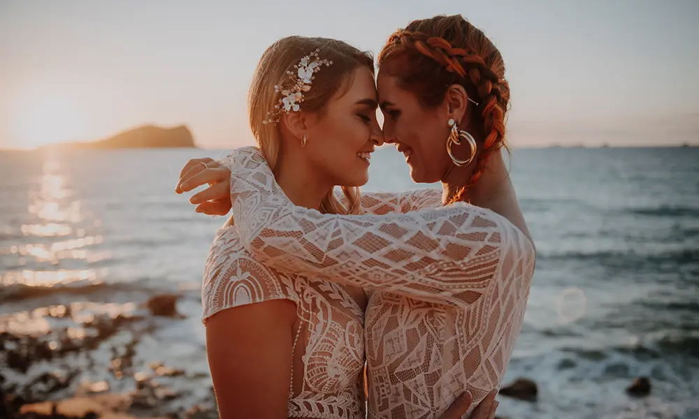 Ein verliebtes Hochzeitspaar am Strand bei Sonnenuntergang: Zwei Bräute in weißen Spitzenkleidern umarmen sich innig, die Sonne taucht das Meer und die Umgebung in ein warmes, goldenes Licht.