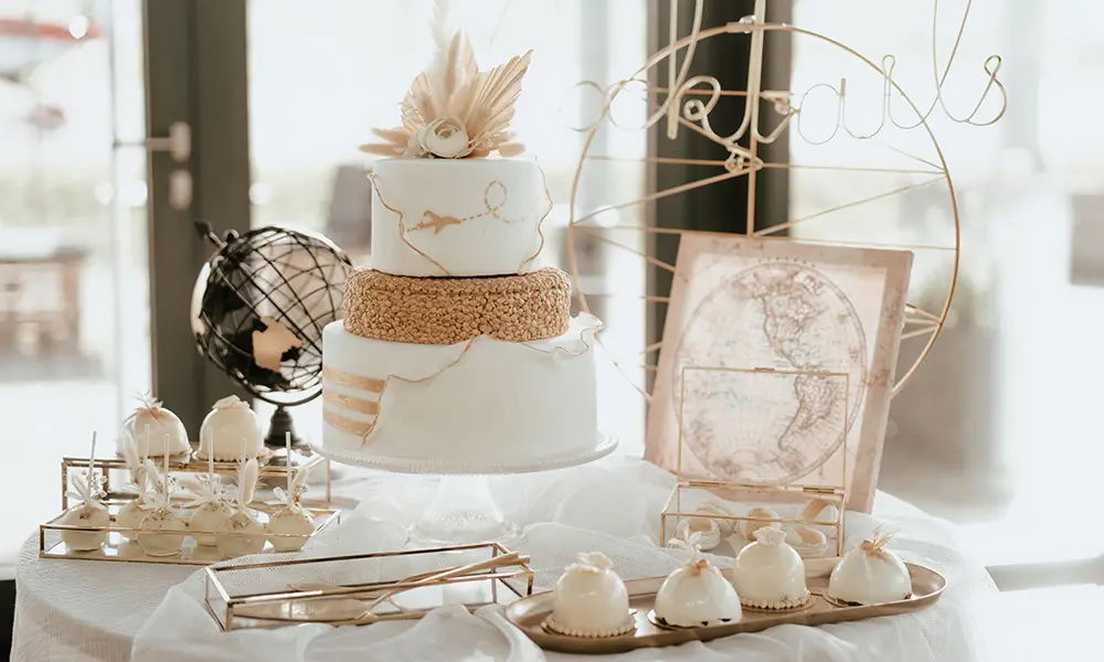Hochzeitstorte mit Weltreise-Thema: Eine zweistöckige weiße Torte mit goldenen Details und einer Karte als Dekoration, umgeben von weißen Cake Pops und kleinen Törtchen, arrangiert auf einem festlichen Tisch.