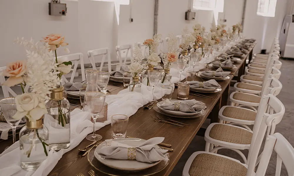 Stilvolle Hochzeitstafel: Eine lange Tafel mit schlichten weißen Stühlen, gedeckt mit grauen Servietten, goldenen Akzenten und weißen Blumen in Glasvasen für eine elegante Dekoration.