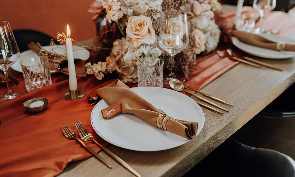 Stimmungsvolle Tischdekoration: Festlich gedeckter Tisch mit einer braunen Stoffserviette, goldenen Details und einer Kerze. Im Hintergrund ein üppiger Blumenstrauß in sanften Creme- und Orangetönen.