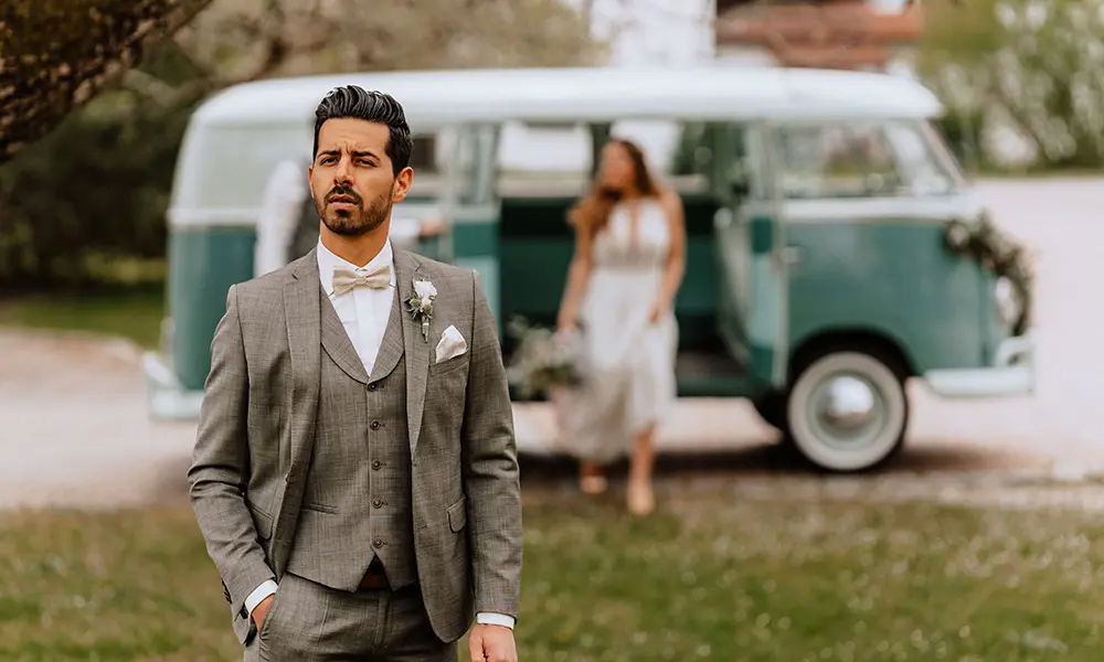 Ein stilvoller Bräutigam in grauem Anzug und Fliege steht selbstbewusst vor einem vintagegrünen VW-Bus, aus dem eine Braut in einem weißen Kleid heraustritt. Der Hintergrund ist leicht unscharf.