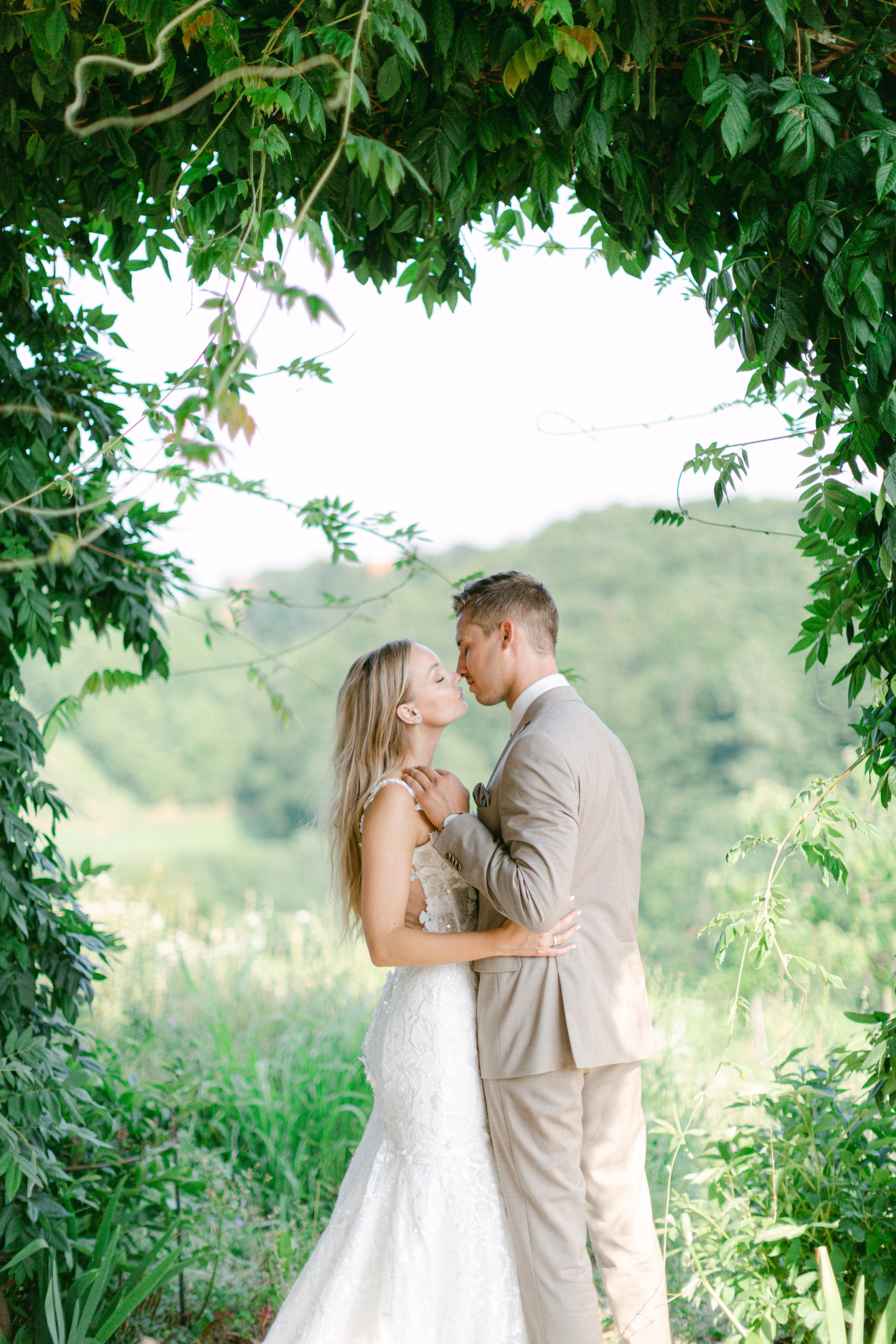 Ein Brautpaar in einem romantischen Moment, umgeben von grünem Laub, das sich für einen Kuss in der Natur neigt.