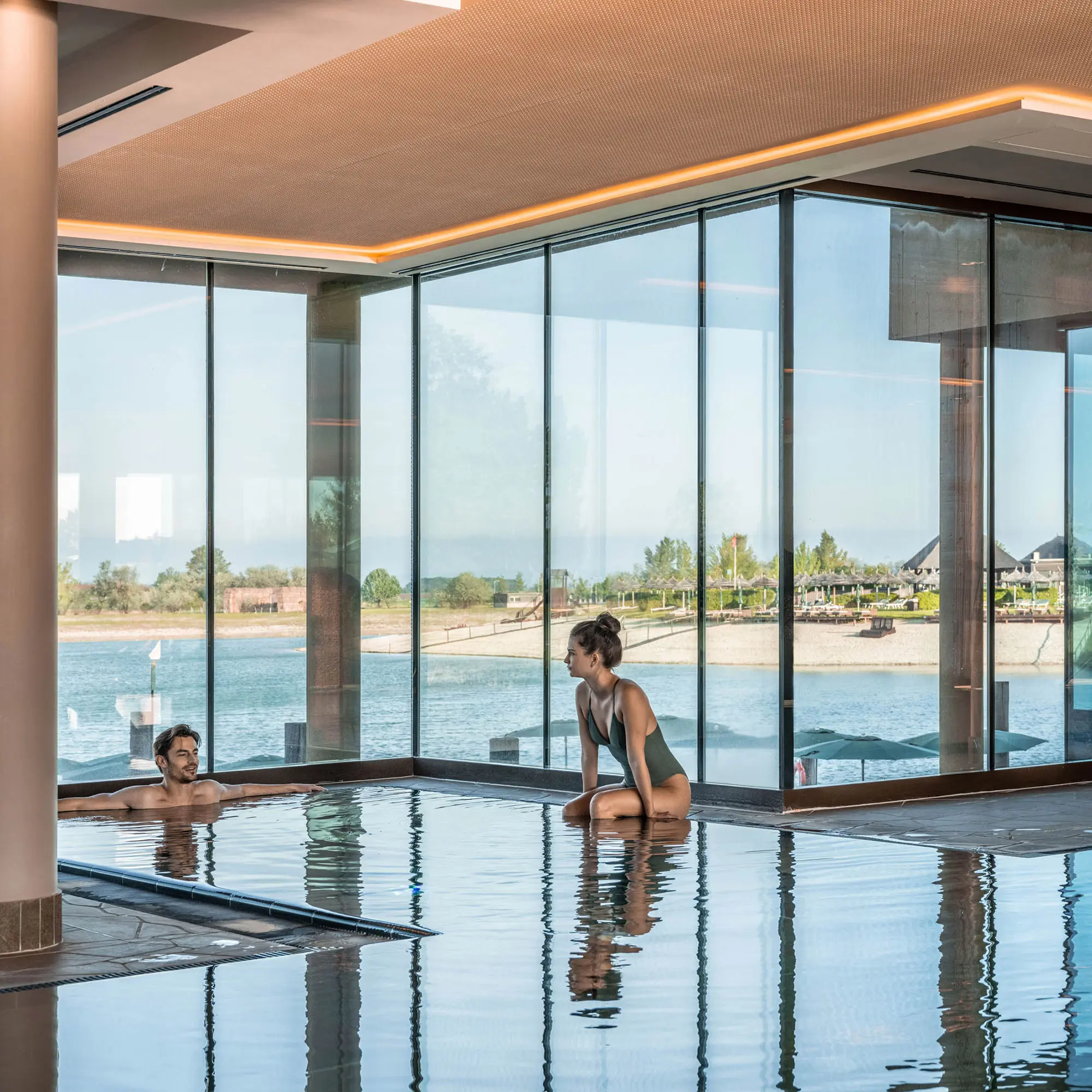 Eine Person sitzt am Rand eines Innenpools und schaut auf einen See, während sie sich mit einer anderen Person unterhält, die im Wasser ist.