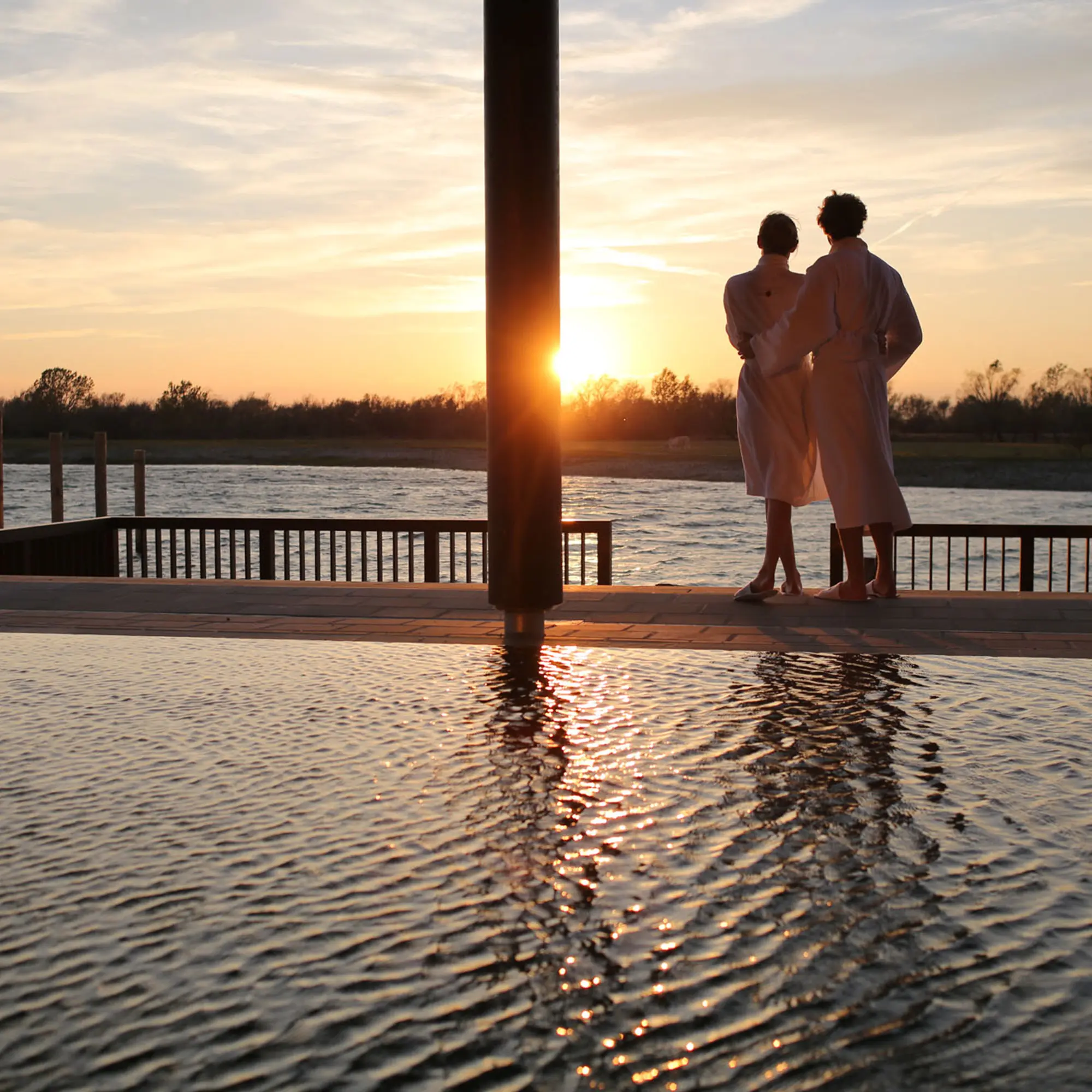 Zwei Personen in Bademänteln genießen den Sonnenuntergang auf einer Anlegestelle, die sich über einen ruhigen See erstreckt.