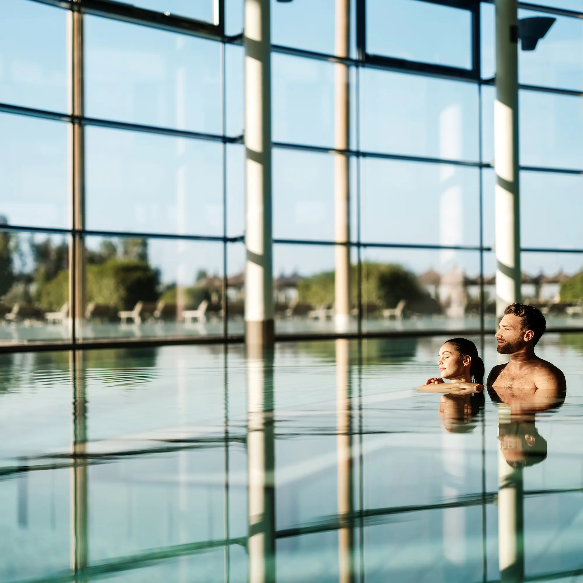Zwei Personen, die in einem Innenpool mit klarem Wasser entspannen, mit einem großen Fenster, das einen Blick auf eine ruhige Wasserlandschaft bietet.