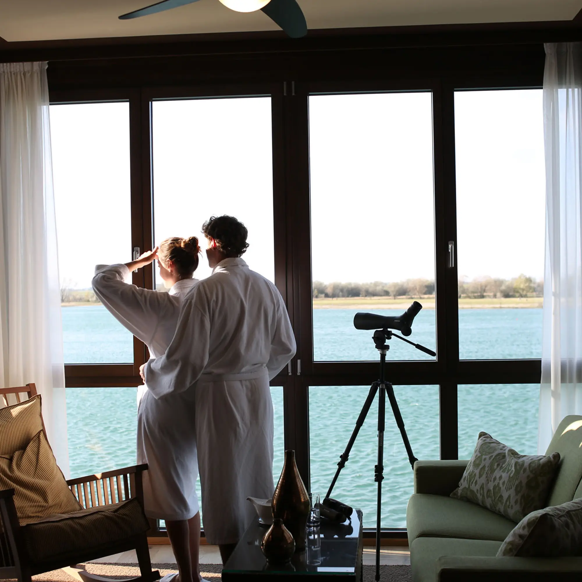 Zwei Personen in weißen Bademänteln stehen vor einem großen Fenster und beobachten die Aussicht auf einen See, neben ihnen steht ein Teleskop.