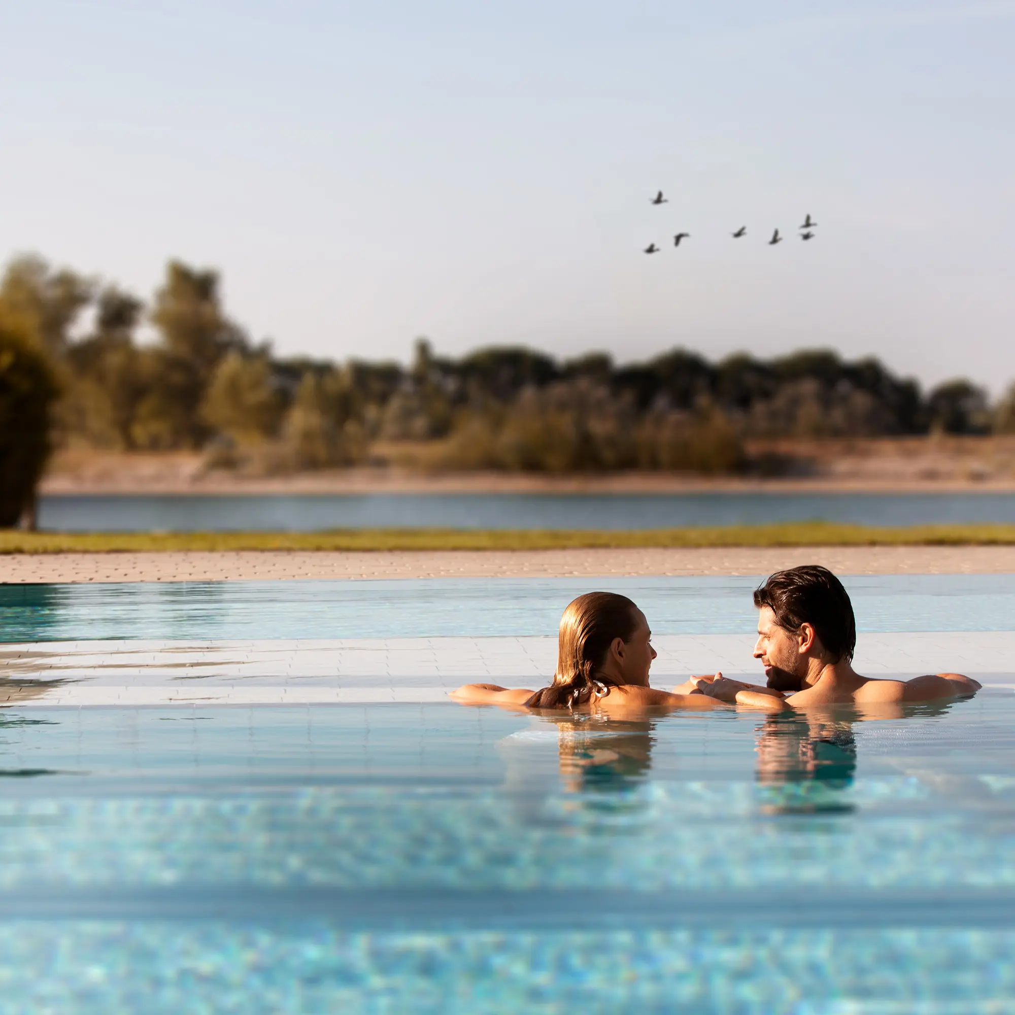 Zwei Personen unterhalten sich im Wasser eines Überlaufpools mit Blick auf eine natürliche Landschaft und fliegende Vögel im Hintergrund.