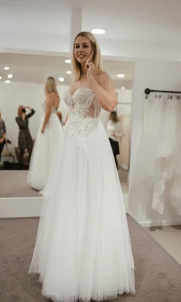 Die zukünftige Braut strahlt in einem trägerlosen Hochzeitskleid mit Spitzenverzierungen bei der Anprobe.