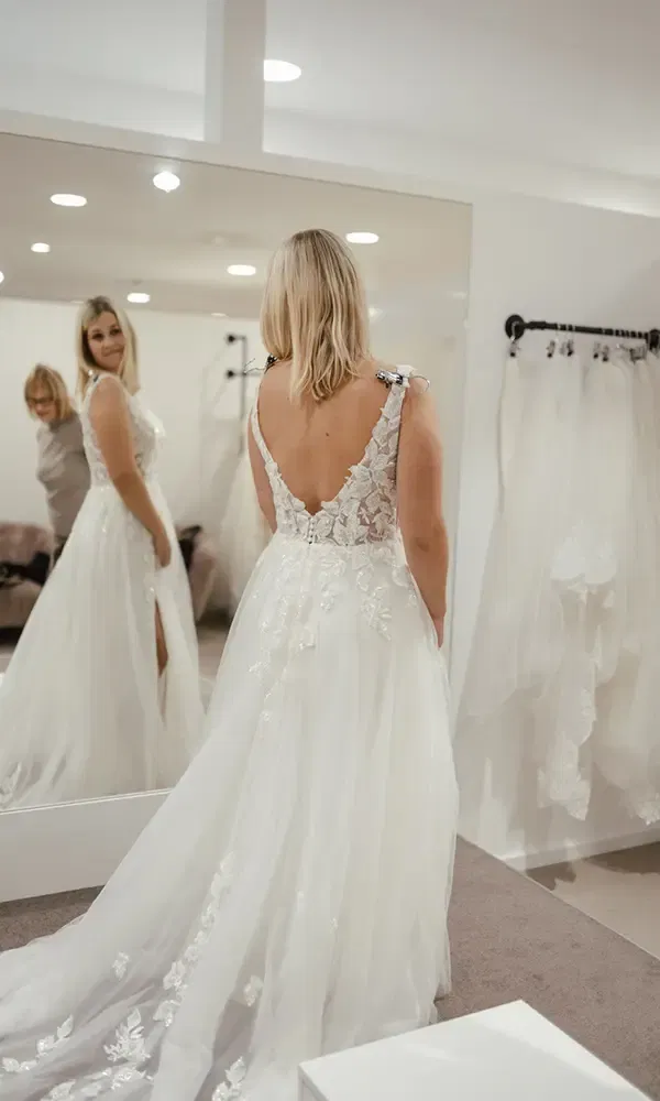 Die Braut steht in einem weißen Kleid vor einem Spiegel und betrachtet sich lächelnd, begleitet von einer Freundin im Hintergrund.