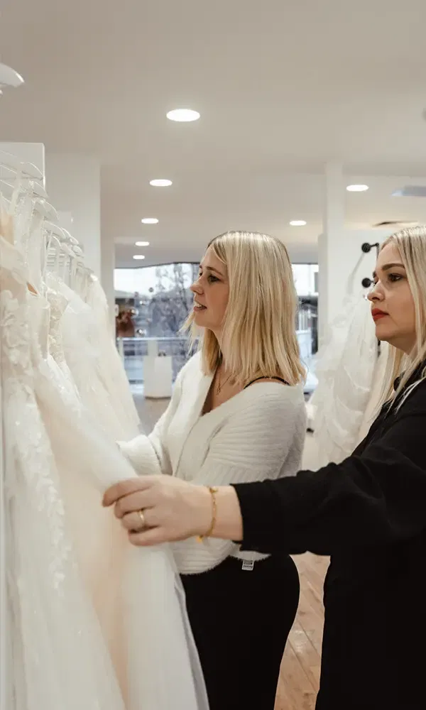 Zwei Frauen durchstöbern eine Auswahl an Hochzeitskleidern in einem eleganten Brautmodengeschäft.