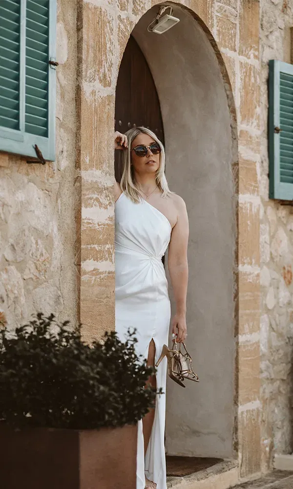 Elegante Braut in einem weißen, schulterfreien Kleid steht lässig in einem rustikalen Torbogen und hält ihre Schuhe in der Hand.