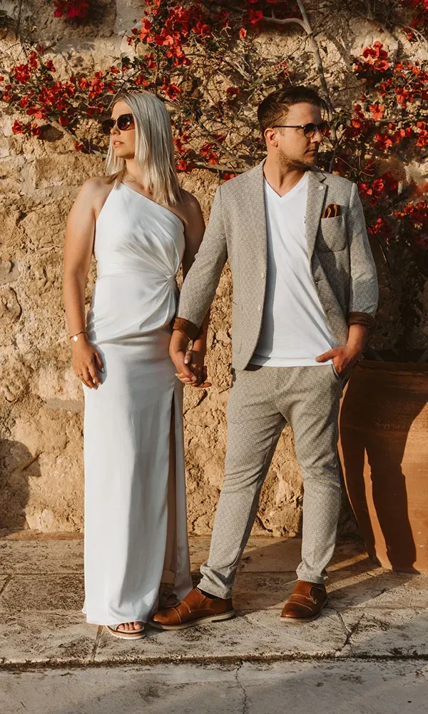 Das Paar steht Hand in Hand vor einer Steinmauer mit roten Blumen im Hintergrund. Die Frau trägt ein weißes Kleid und eine Sonnenbrille, der Mann einen grauen Anzug und ein weißes T-Shirt.
