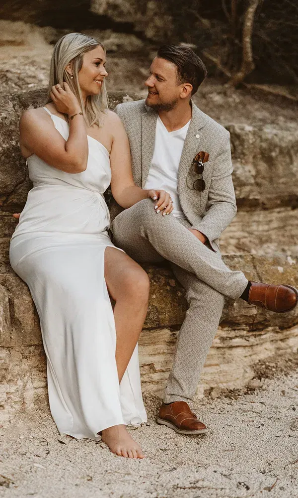 Verliebtes Paar in stilvollen Outfits sitzt lächelnd auf einem Felsen; sie in einem weißen Kleid, er im hellgrauen Anzug.