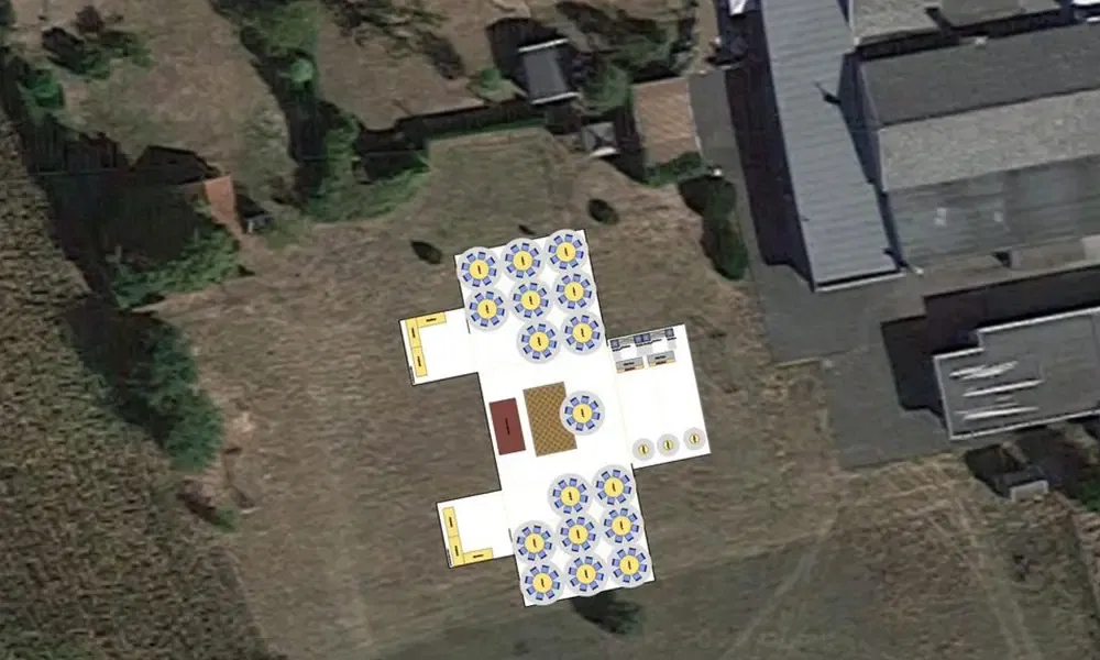 Luftaufnahme eines großen Festzeltes mit runden Tischen und Stühlen, das auf einer Wiese neben Gebäuden aufgestellt ist. Die Zeltanordnung zeigt mehrere Tische mit gelb-blauen Stühlen.