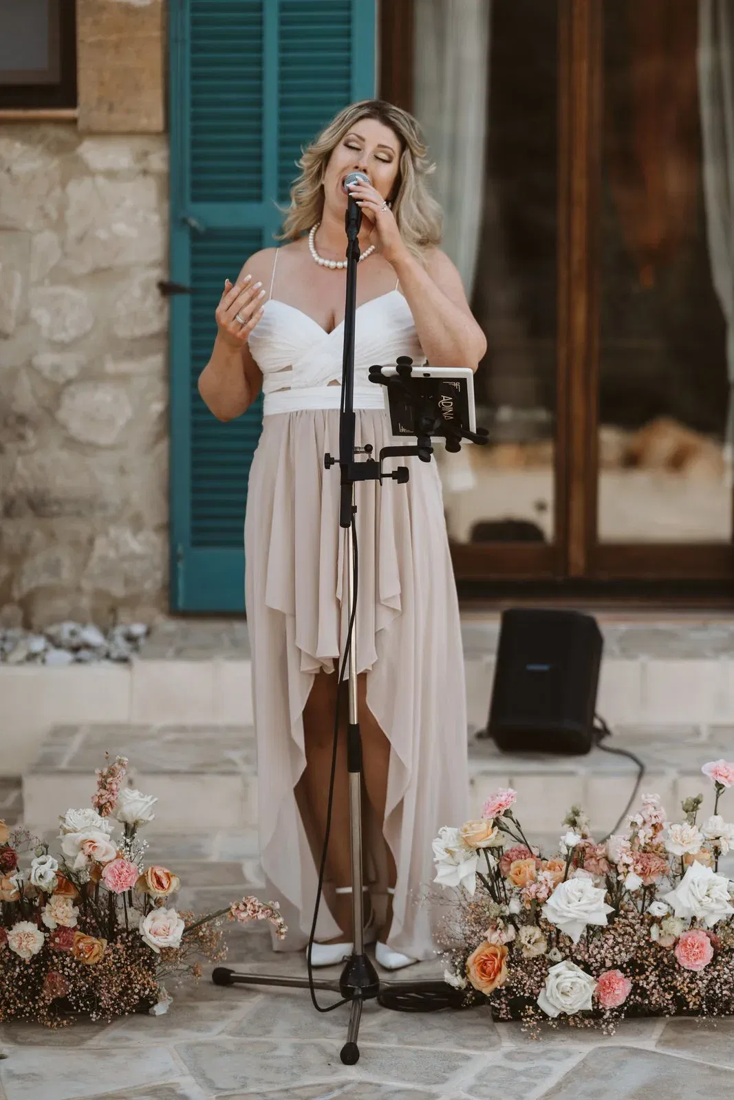 Sängerin im cremefarbenen Kleid singt während einer Hochzeit neben blumigen Dekorationen, ein emotionaler Moment für die Gäste.