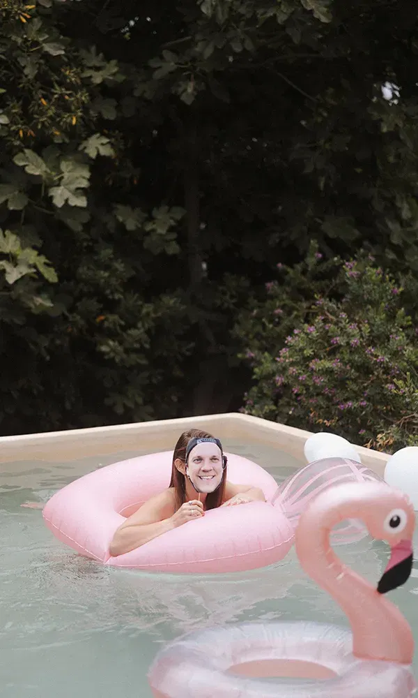Eine Person entspannt sich auf einem rosa Schwimmring im Pool und trägt eine Maske mit dem Gesicht eines Mannes. Neben ihr schwimmt ein aufblasbarer Flamingo.