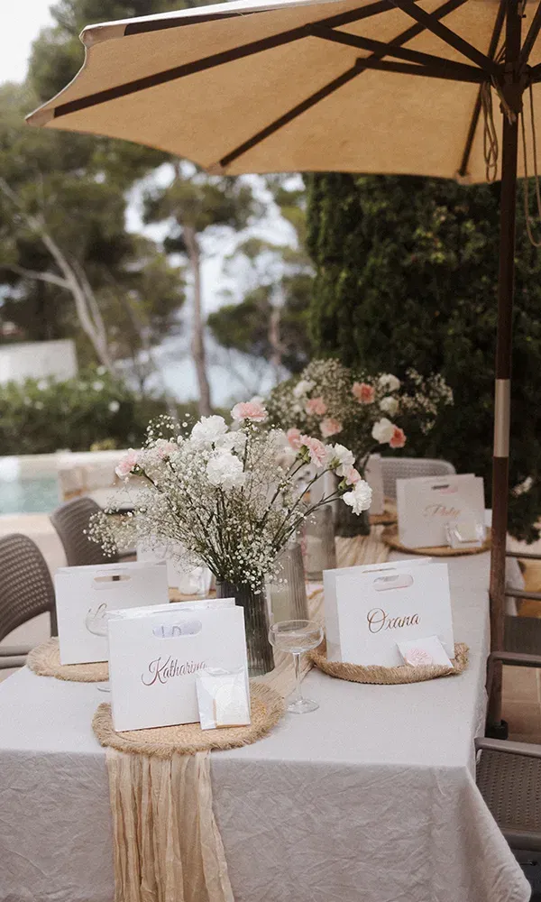 Ein festlich gedeckter Tisch im Freien mit personalisierten Geschenktüten und Blumenarrangements. Auf dem Tisch stehen Vasen mit rosa und weißen Blüten unter einem großen Sonnenschirm.