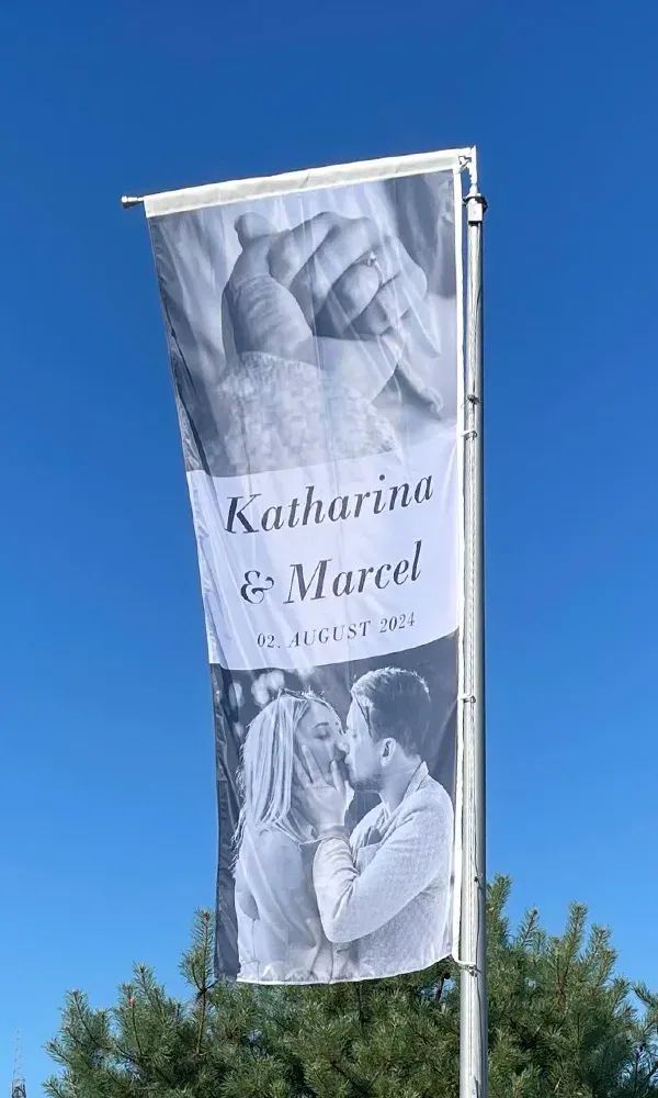 Eine große Flagge mit einem Schwarz-Weiß-Foto des Brautpaares Katharina und Marcel, mit ihrem Hochzeitsdatum „02. August 2024“, weht vor einem klaren, blauen Himmel.