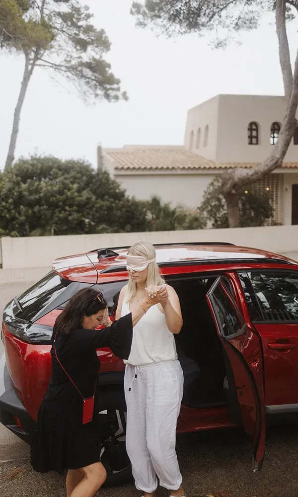 Eine Frau mit verbundenen Augen wird von einer Freundin aus einem roten Auto geführt. Im Hintergrund ist ein Haus und grüne Bäume zu sehen.