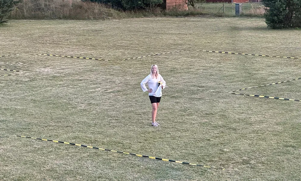 Frau steht auf einer Wiese, die mit gelb-schwarzem Absperrband markiert ist, lächelnd in die Kamera schauend. Die markierten Flächen zeigen den geplanten Aufbaubereich für ein Event.