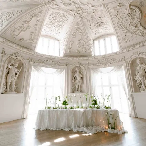 Elegant gedeckter Hochzeitstisch mit weißer Tischwäsche, zarten Blumenarrangements und Kerzen, inszeniert in einem prunkvollen Palais mit reich verzierten Stuckdecken, Skulpturen und großen Fenstern, die den Raum mit Tageslicht fluten.