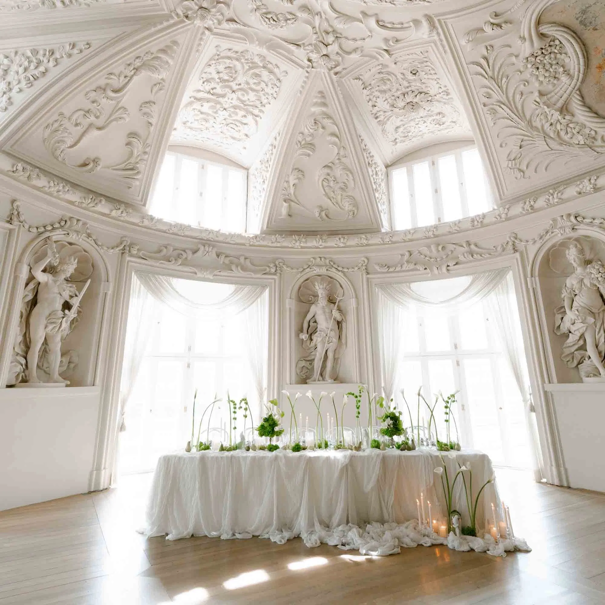 Elegant gedeckter Hochzeitstisch mit weißer Tischwäsche, zarten Blumenarrangements und Kerzen, inszeniert in einem prunkvollen Palais mit reich verzierten Stuckdecken, Skulpturen und großen Fenstern, die den Raum mit Tageslicht fluten.