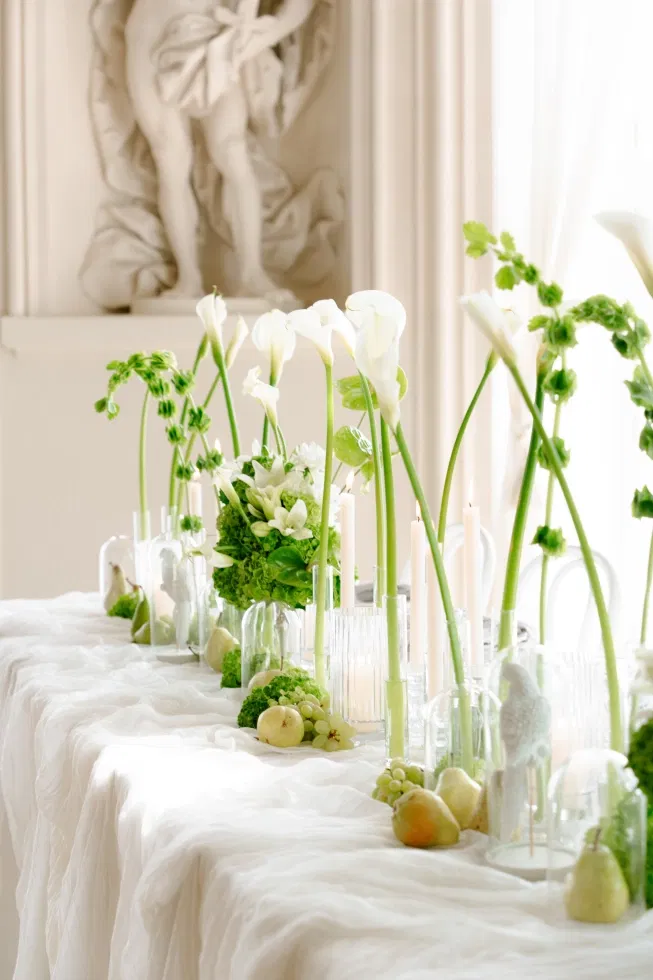 Elegante Hochzeitstafel mit weißen Calla-Lilien, grünen Blüten, Kerzen in Glasvasen und dekorativen Früchten auf fließendem Stoff.
