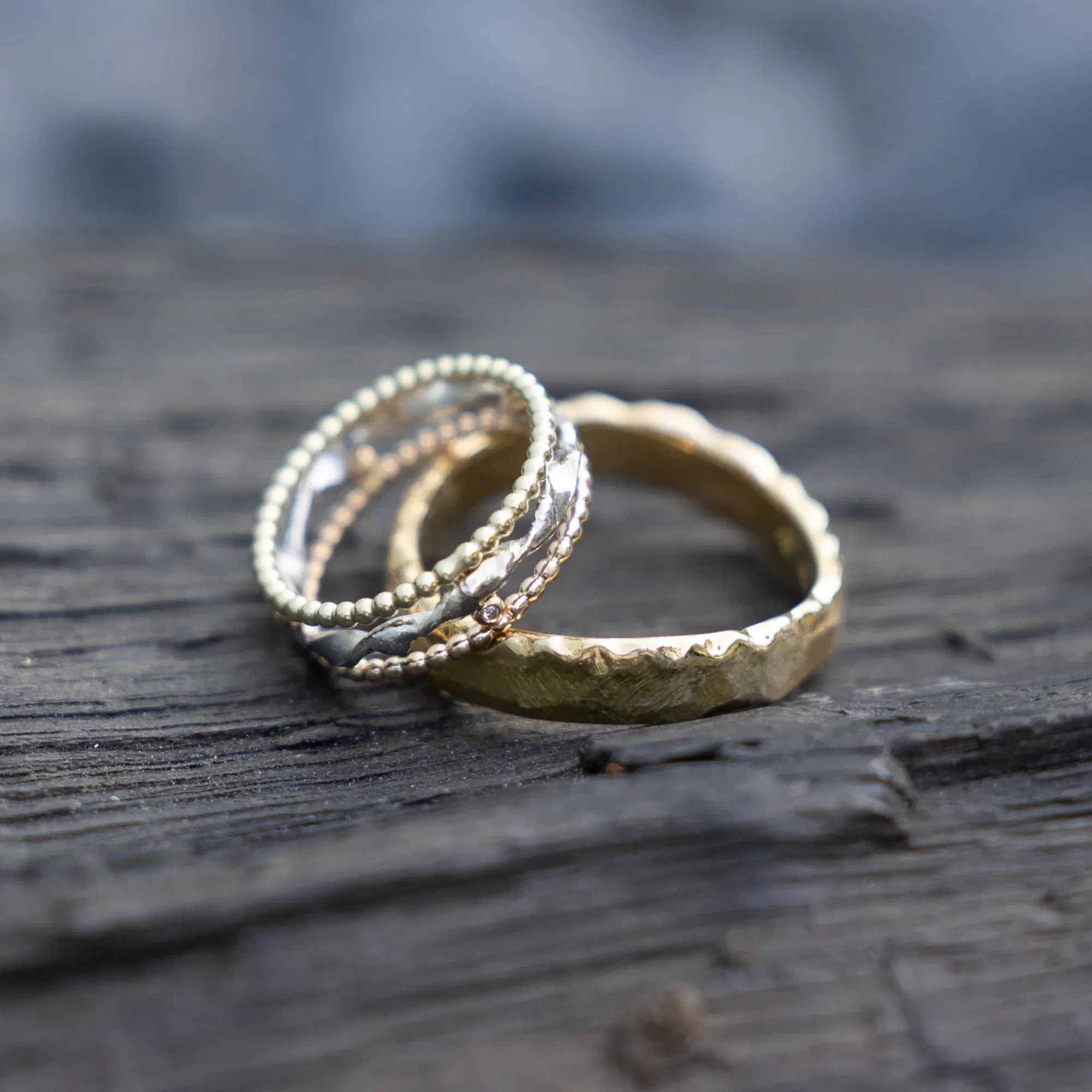 Mehrere filigrane Trauringe in Gold- und Silberfarben, übereinandergestapelt auf einer Holzoberfläche, kombiniert aus verschiedenen Materialien und Designs.