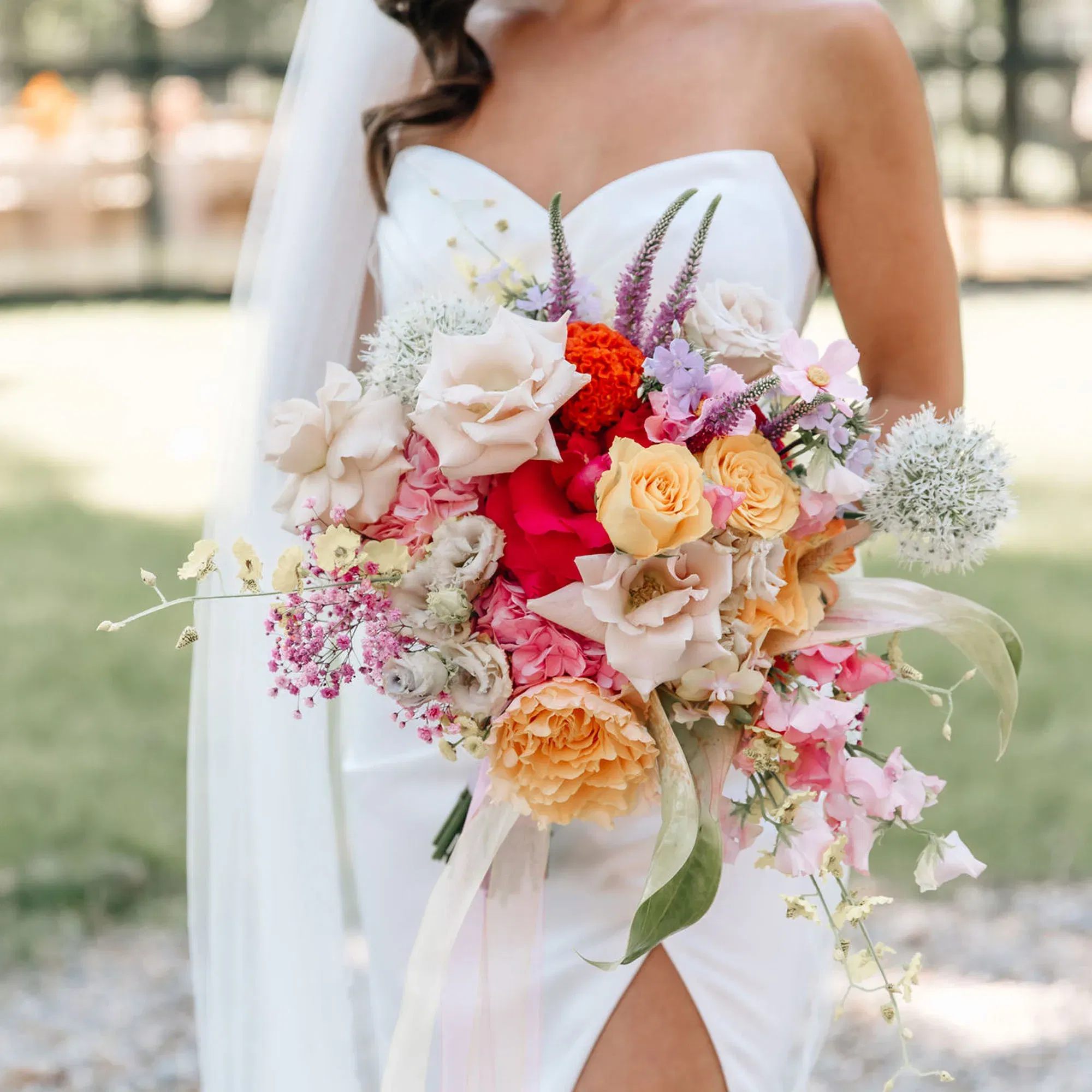 Nahaufnahme des Oberkörpers einer Braut in einem schlichten weißen Kleid. Sie trägt einen üppigen, farbenfrohen Blumenstrauß mit orangen, pinken und cremefarbenen Blüten sowie lila Akzenten. Die Umgebung ist grün und leicht verschwommen.