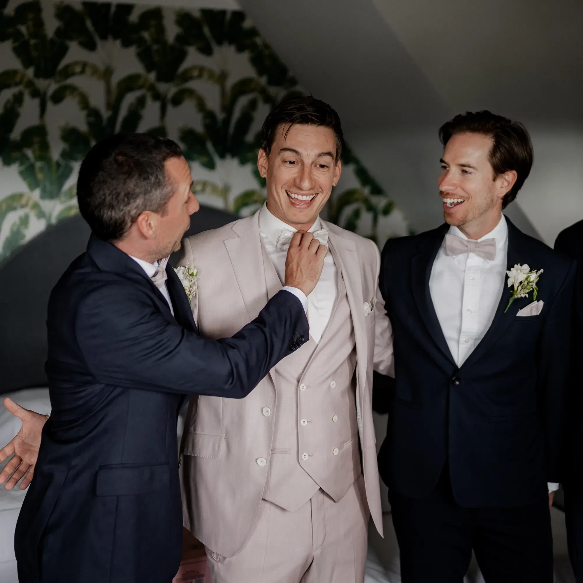 Eine Gruppe von drei Männern, darunter der Bräutigam in einem beigen Anzug, lachen gemeinsam, während ein Groomsman seine Fliege richtet.